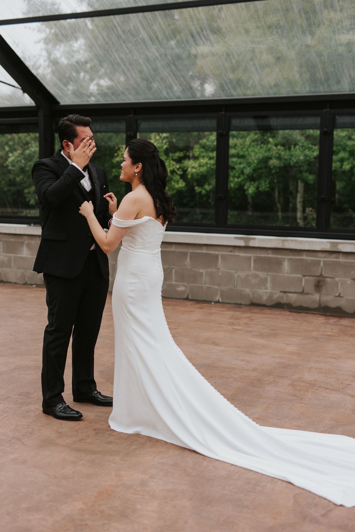 An emotional first look between newlyweds in formal attire under a glass ceiling on a rainy day.