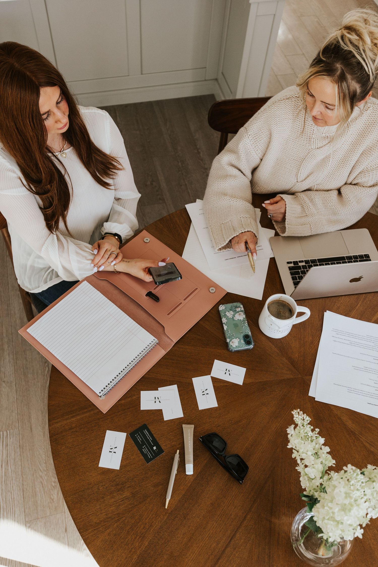 Two women working together at wooden table with papers, laptop and white flowers in home office setting.