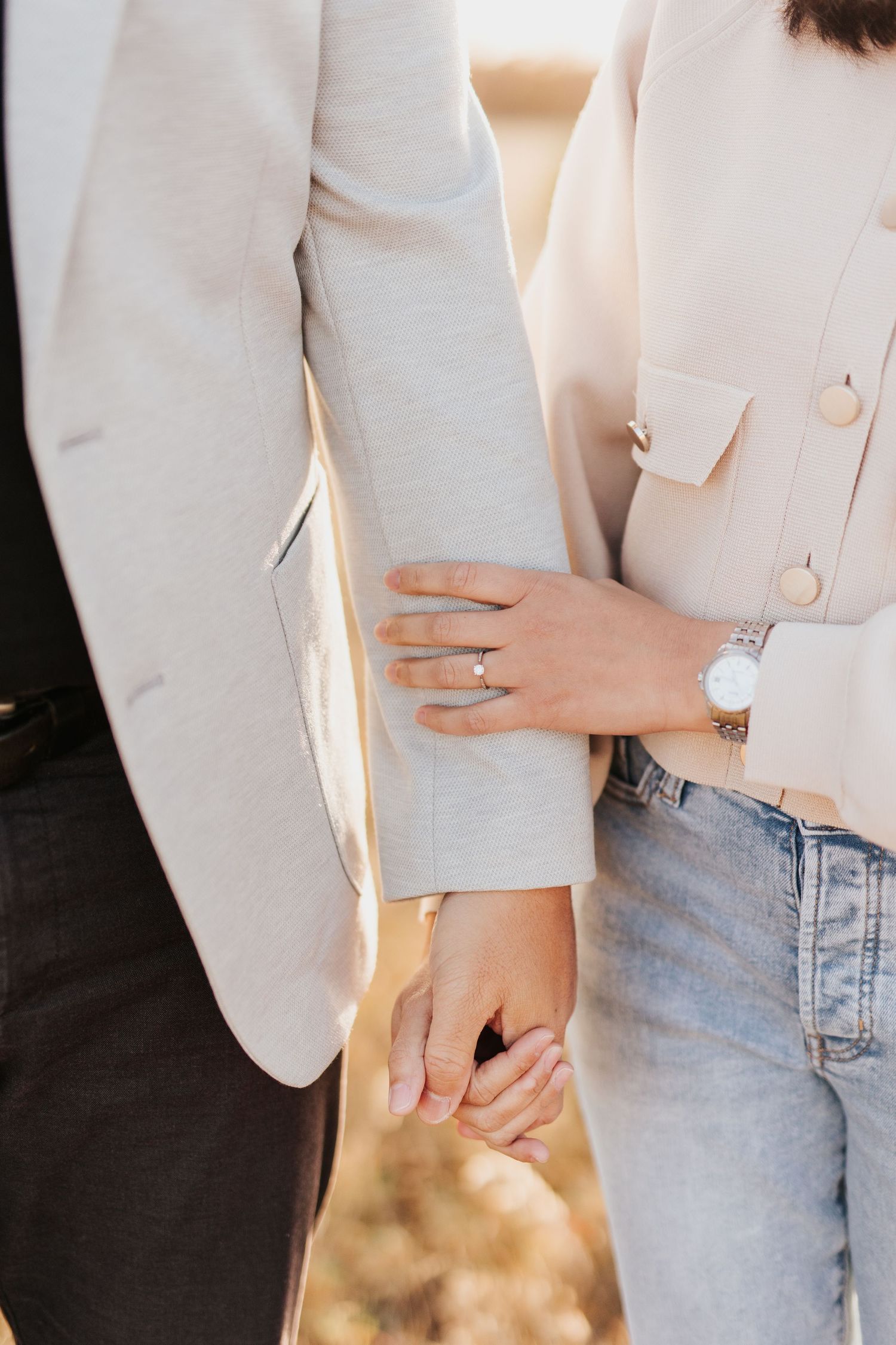 Close-up detail of couple holding hands with engagement ring visible on light cream blazer background.