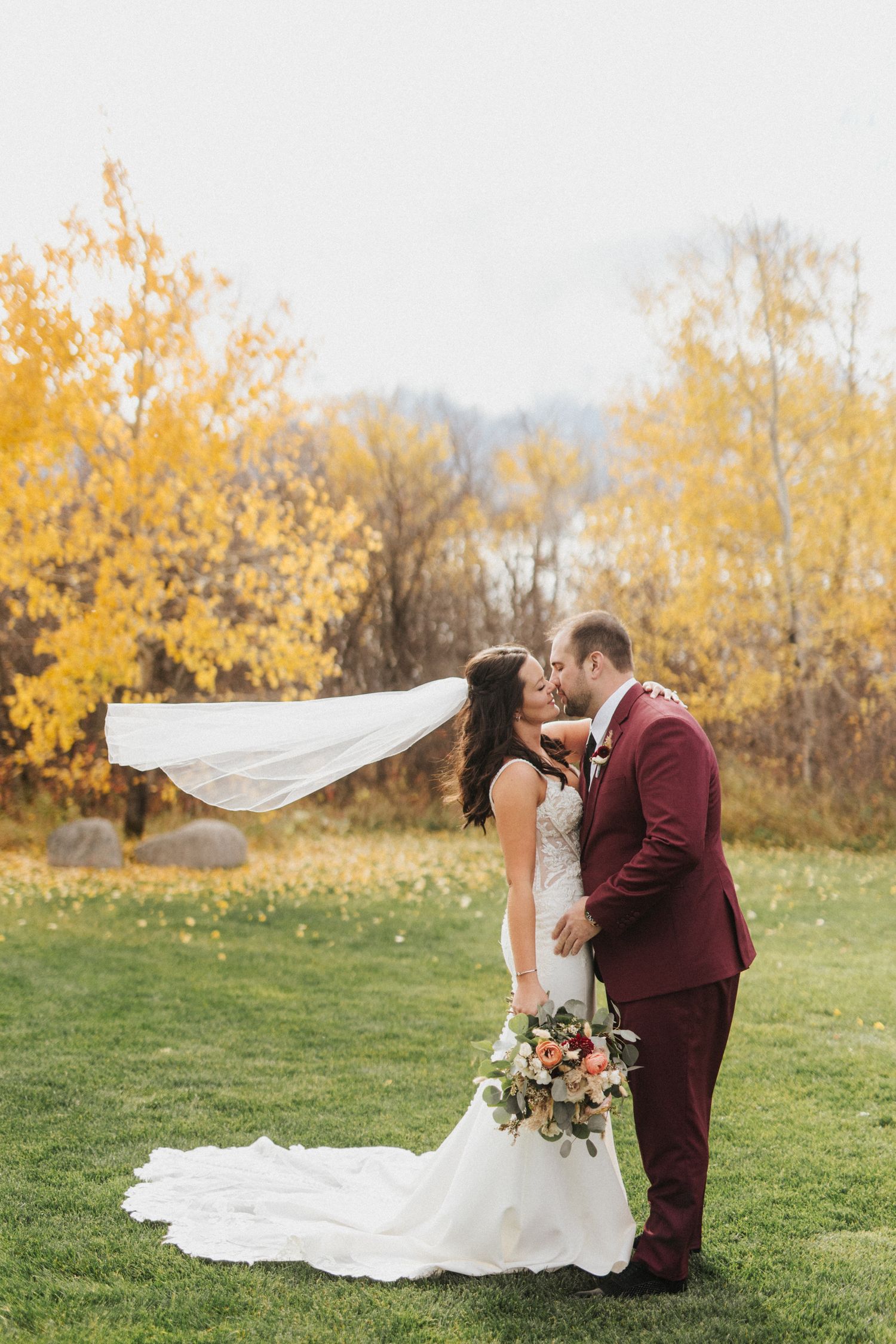 A couple embraces on a grassy field with vibrant yellow autumn trees in the background.