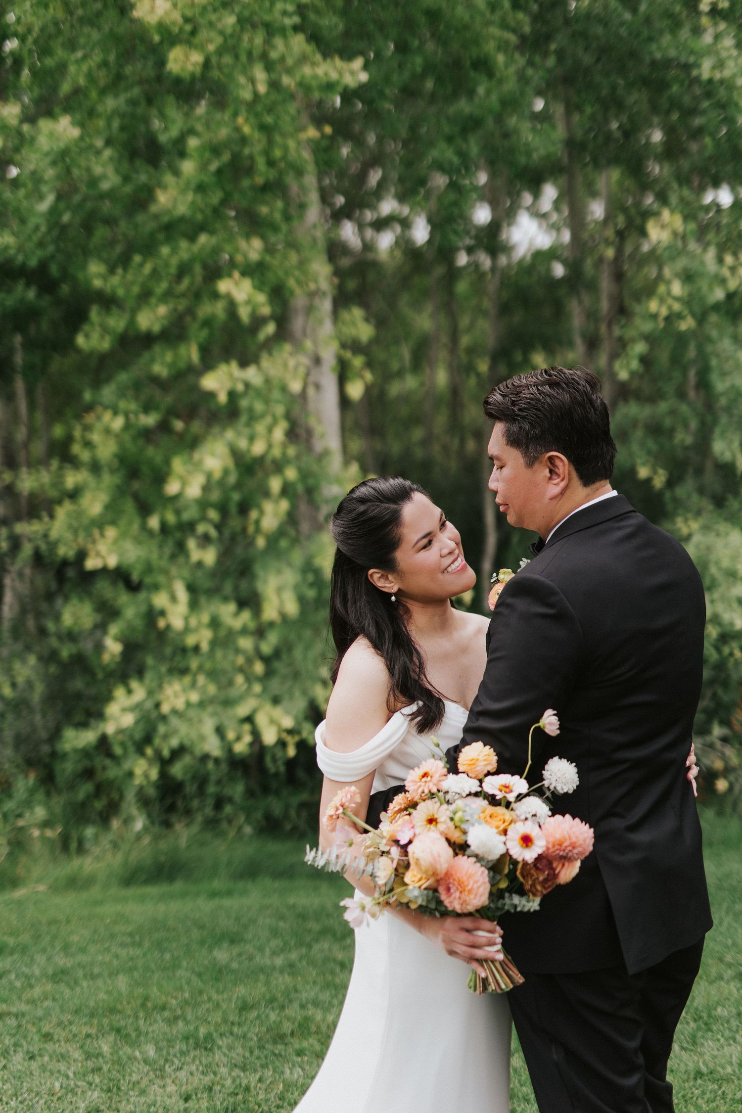 A wedding couple embraces outdoors holding a vibrant peach and coral floral bouquet.