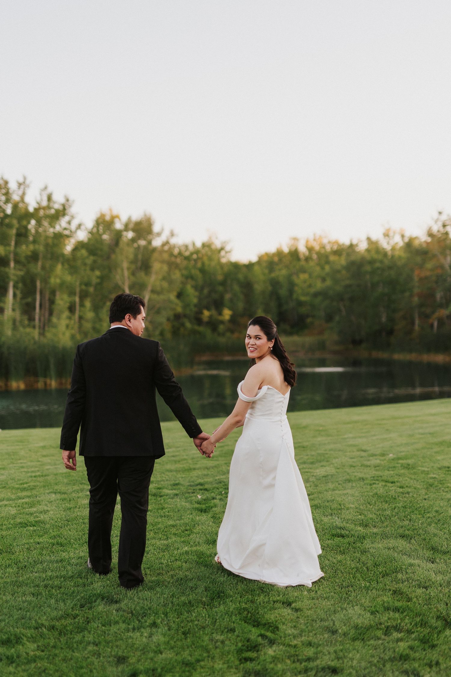 Newlyweds walk hand in hand on grass beside a serene lake during golden hour.