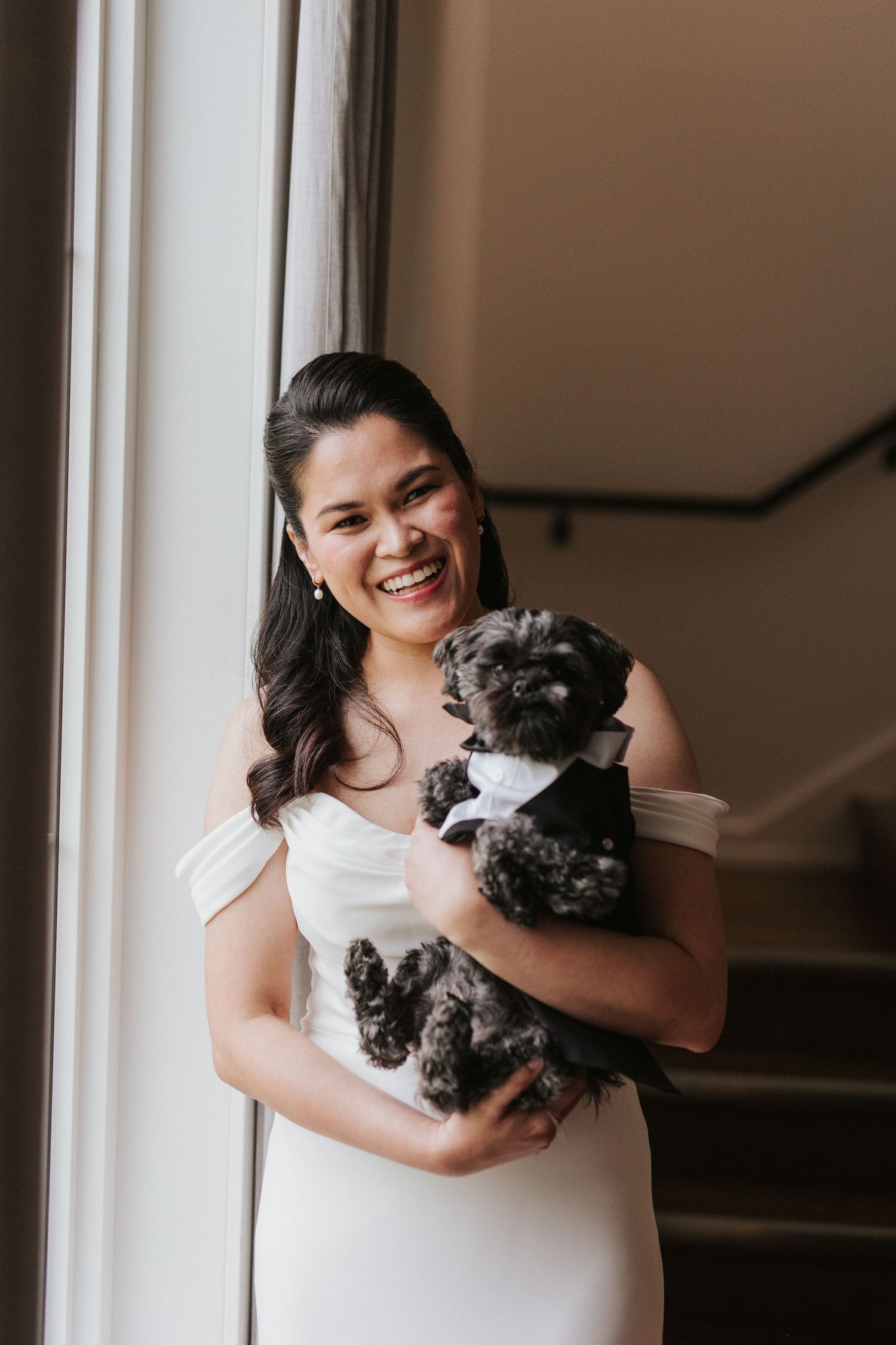 A bride in a white dress joyfully embraces a small black dog in an indoor setting.