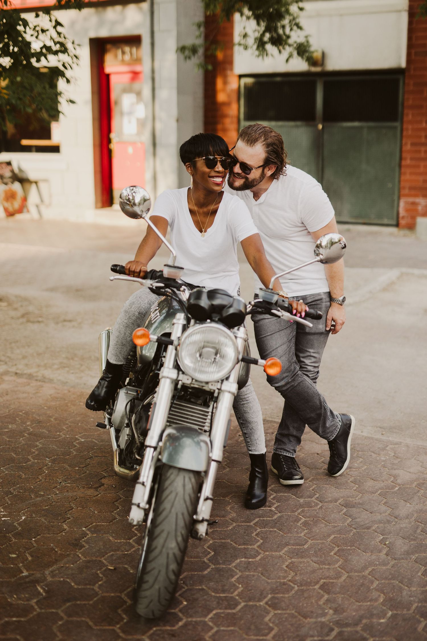 Playful couple posing with a motorcycle on urban cobblestone street.