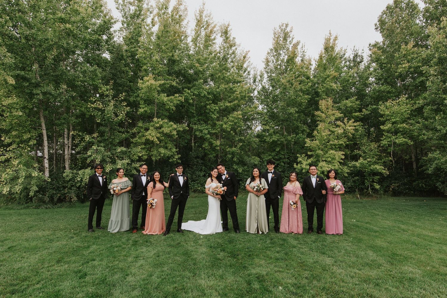 Wedding party in formal attire stands on green lawn with forest backdrop during outdoor ceremony.