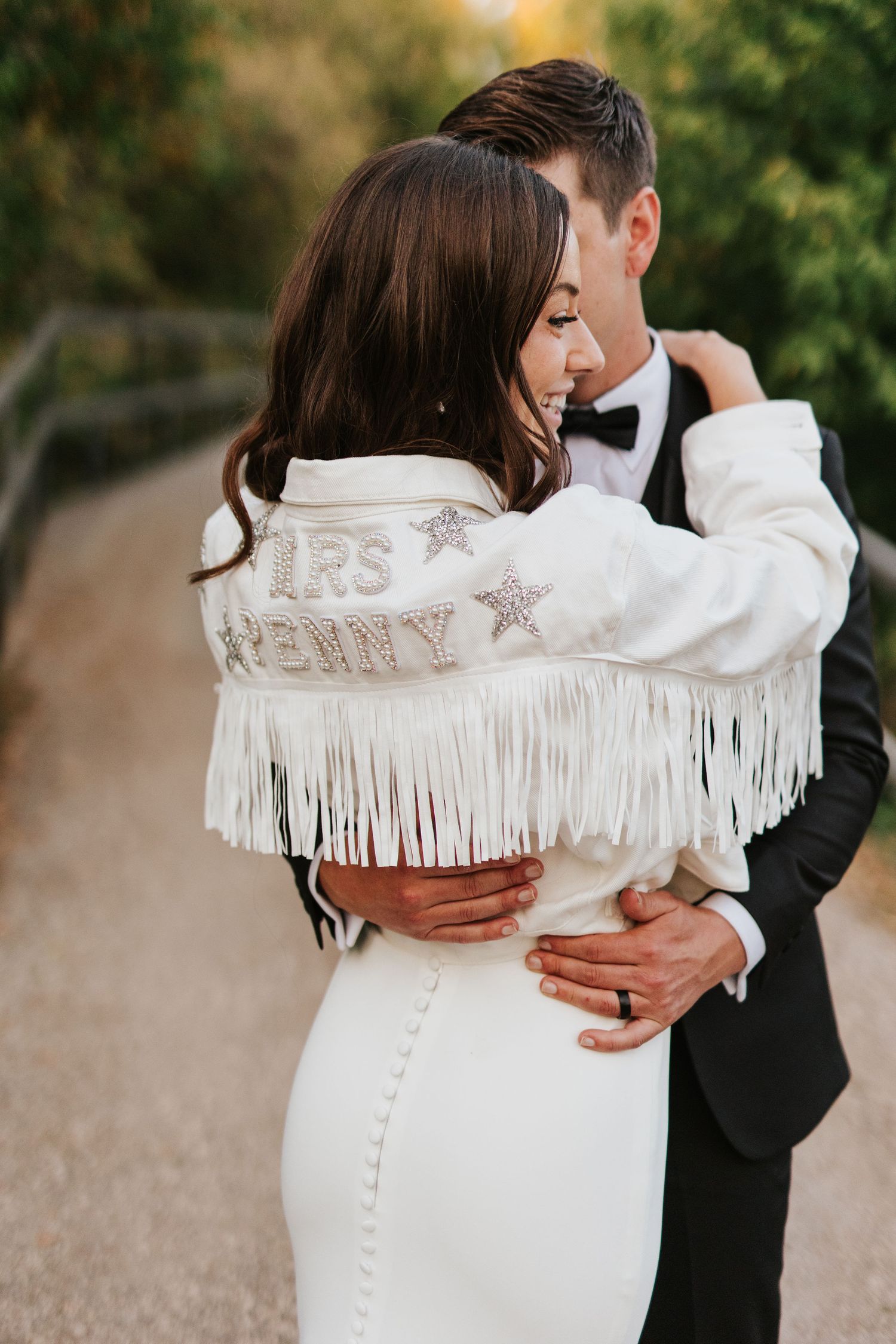 Back view of newlyweds with decorative fringe jacket against autumn foliage.
