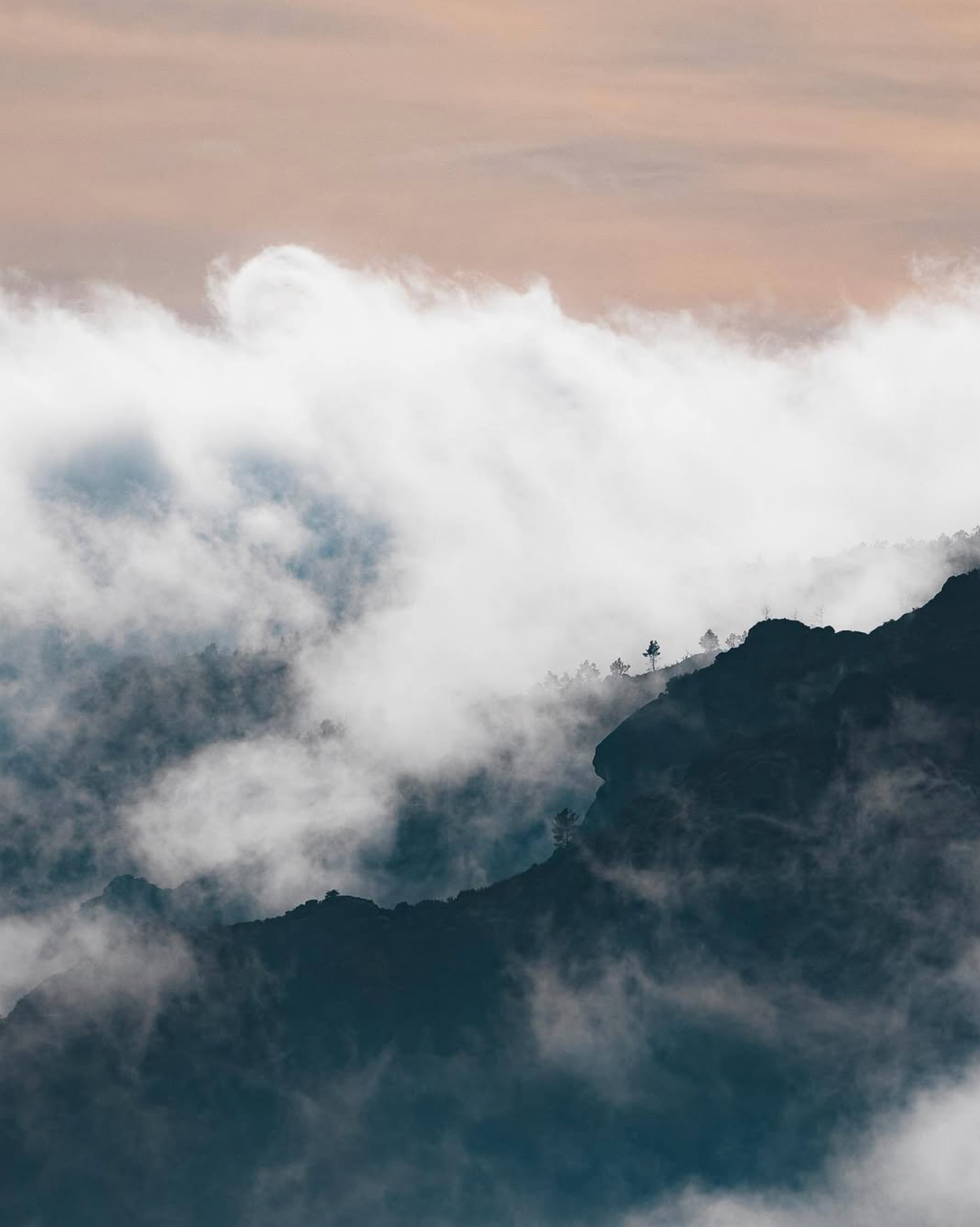 Misty mountain peaks shrouded in dramatic clouds and fog creating a moody atmospheric landscape at sunset.