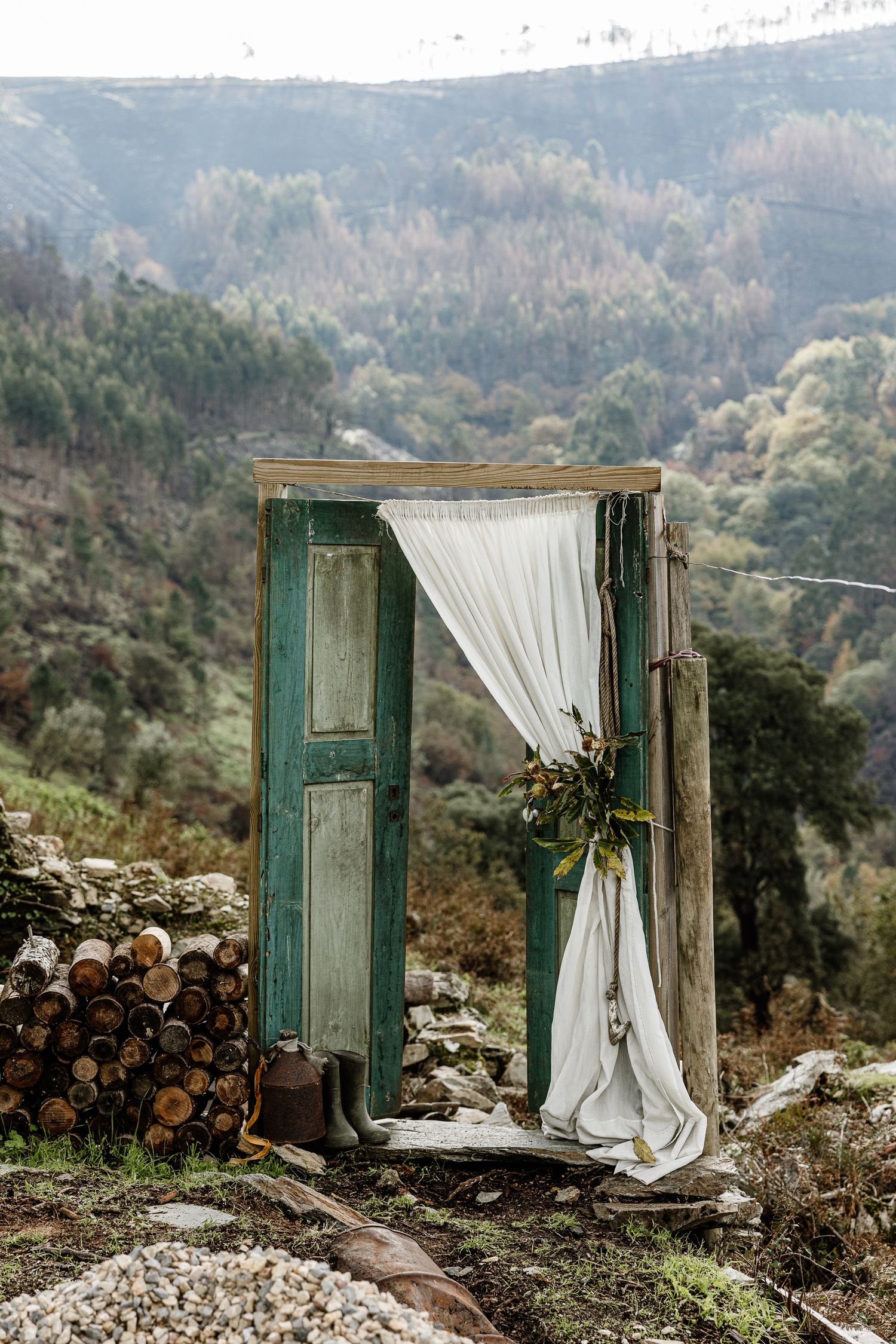 A vintage green door with white curtain stands alone on a mountainside with stacked firewood and scenic forest views.