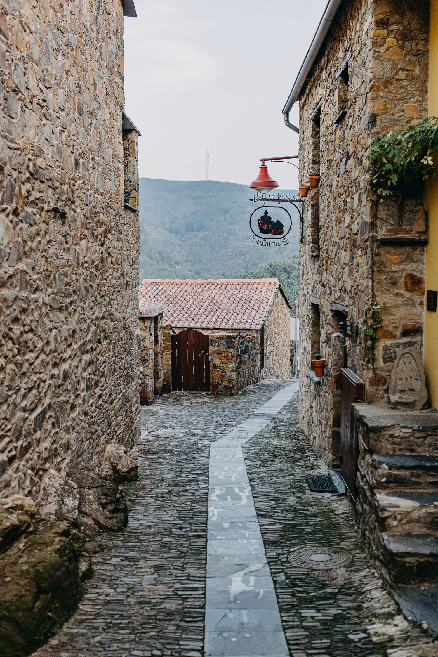 Narrow cobblestone alley winding through ancient stone buildings in a medieval European village with mountain views.