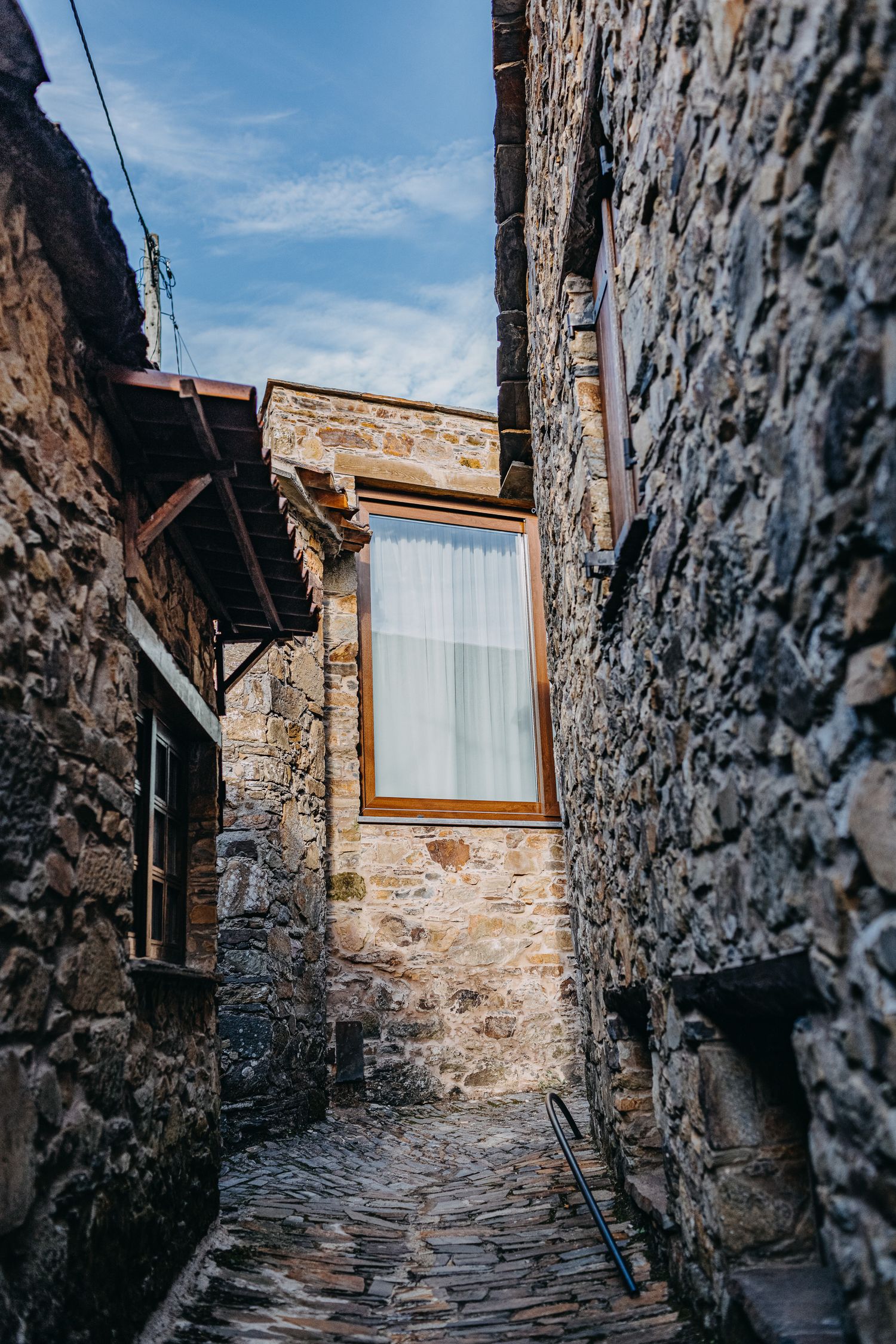 Narrow stone alley with rustic walls and a window in an ancient village leads to blue sky above.