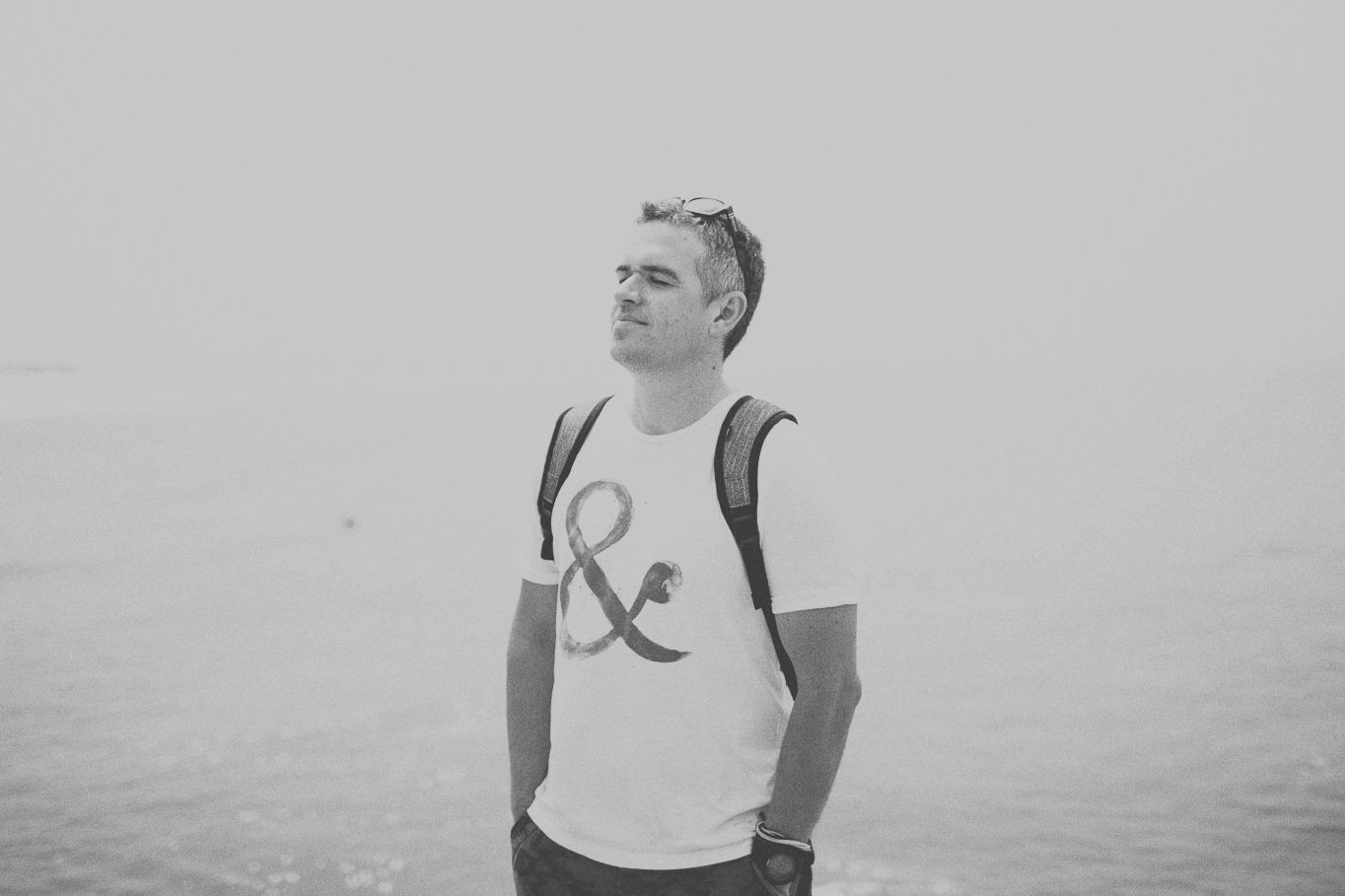 A person in a white ampersand t-shirt and backpack stands against a foggy outdoor background in black and white.