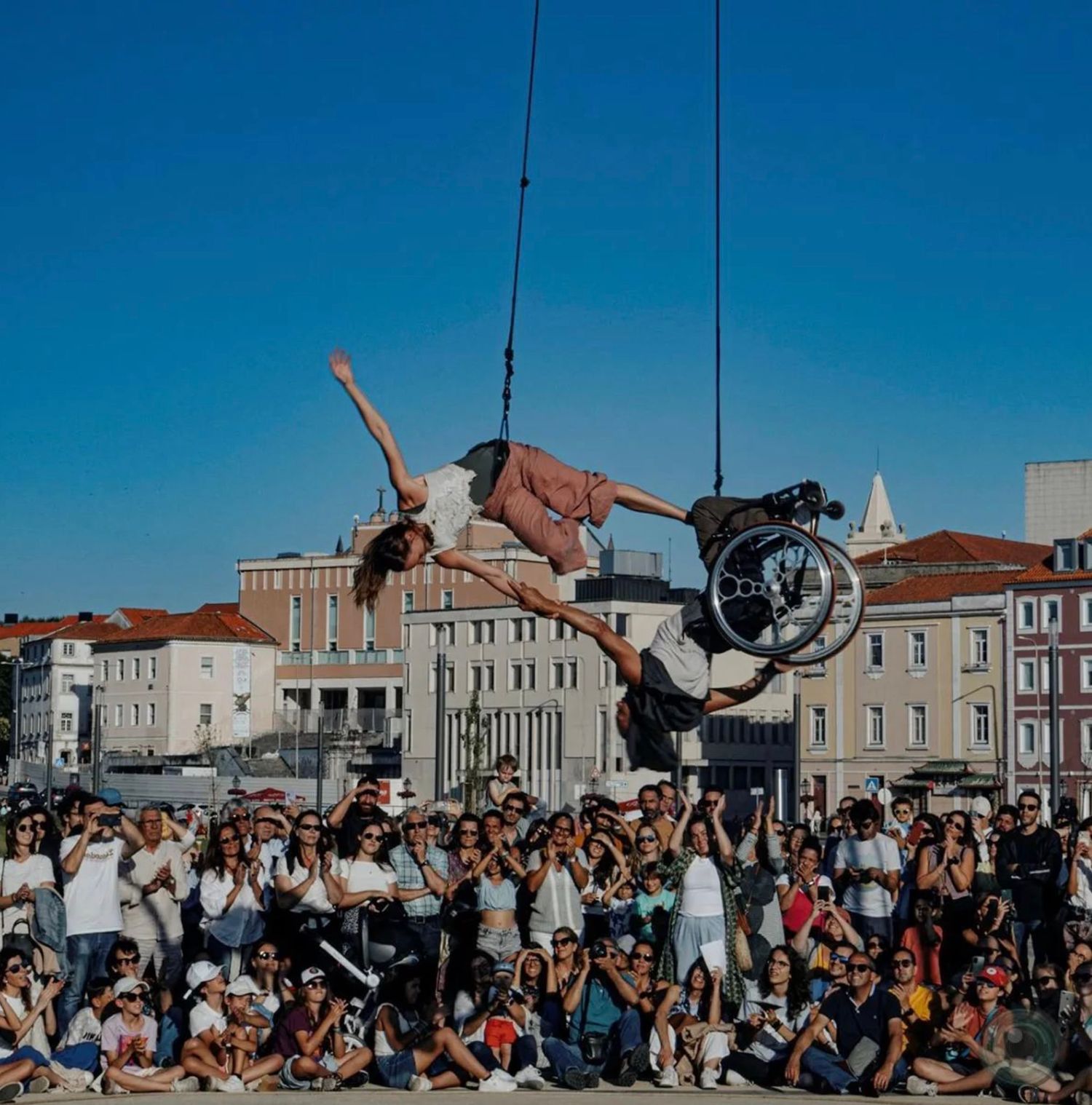 Street performer suspended with wheelchair doing an acrobatic display in front of large crowd in city square.