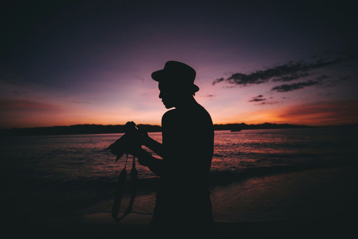 A silhouetted figure stands against a dramatic purple sunset sky while taking photographs at the ocean's edge.