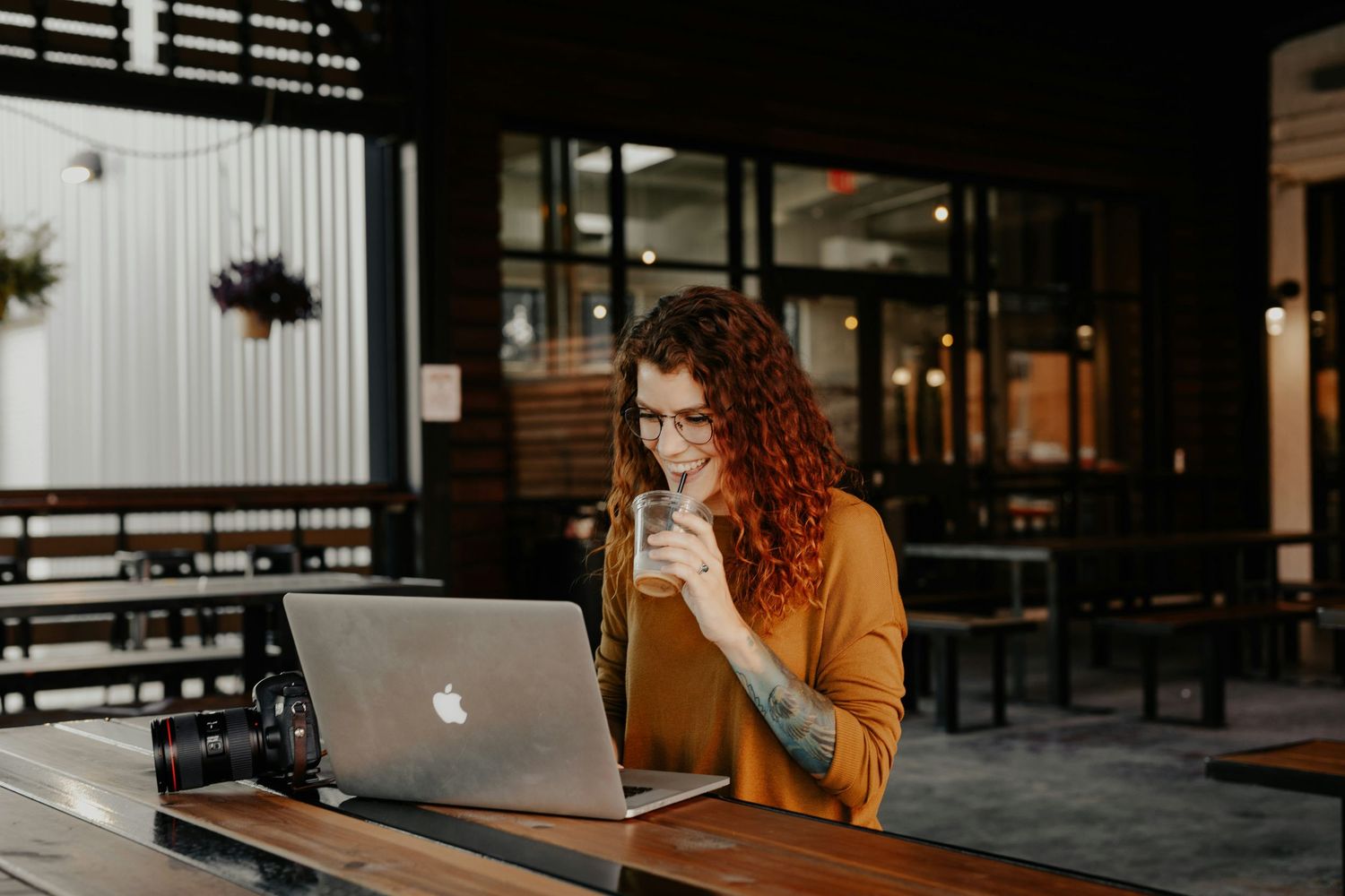 A person in a brown sweater works on a laptop at a cafe table near large windows during golden hour.