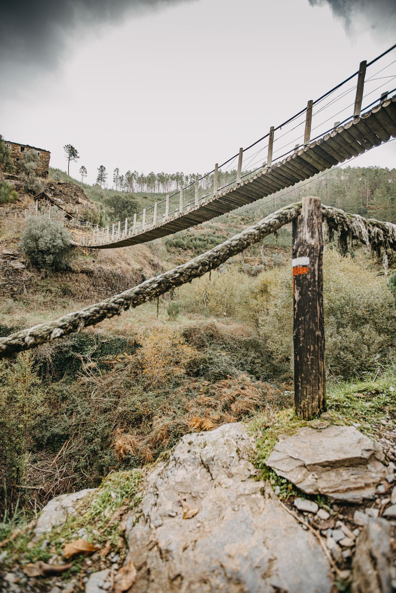 A rustic wooden suspension bridge stretches across a rocky ravine in a mountainous landscape.