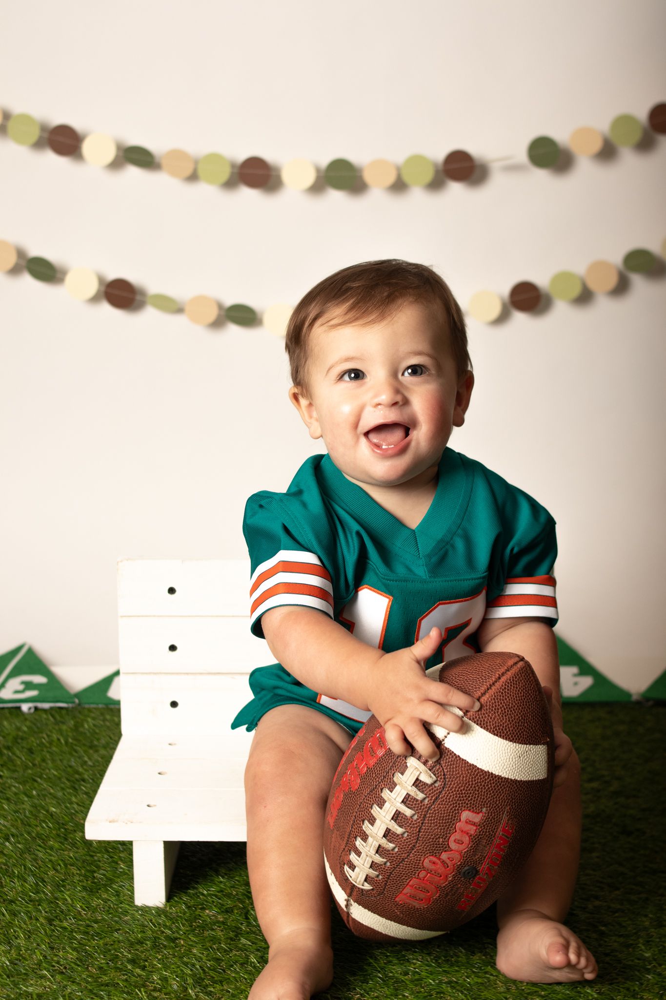Happy baby in teal football jersey sitting on bench with football.