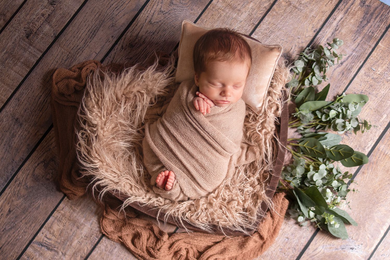 Sleeping newborn wrapped in beige cloth on fur blanket with greenery accents on wooden floor.