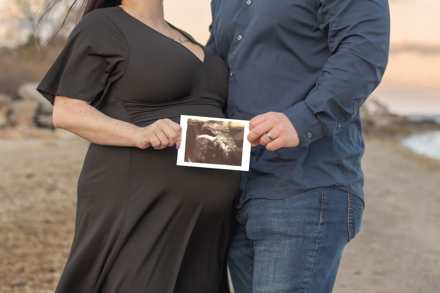 A pregnant woman in black dress and partner holding ultrasound photo outdoors at sunset at Rocky Point Beach Warwick RI