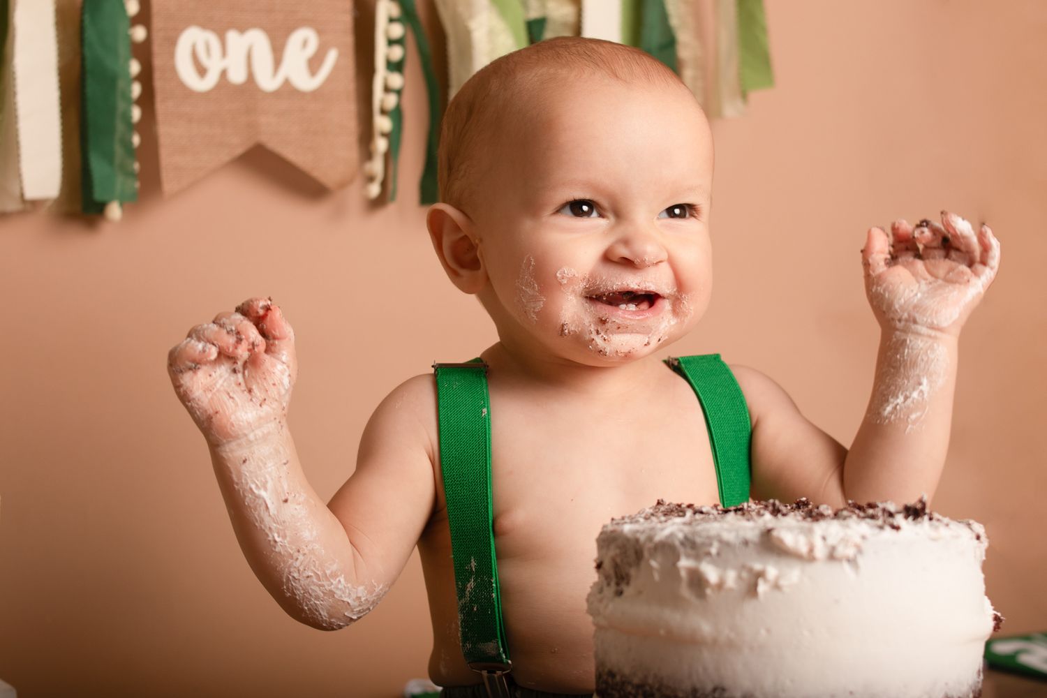 Messy cake smash photo with happy baby wearing green suspenders.