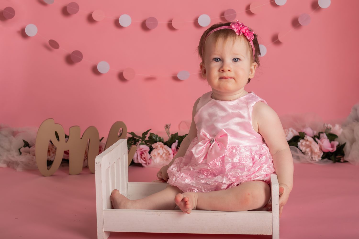 Baby in pink dress sitting on small white chair against pink backdrop.