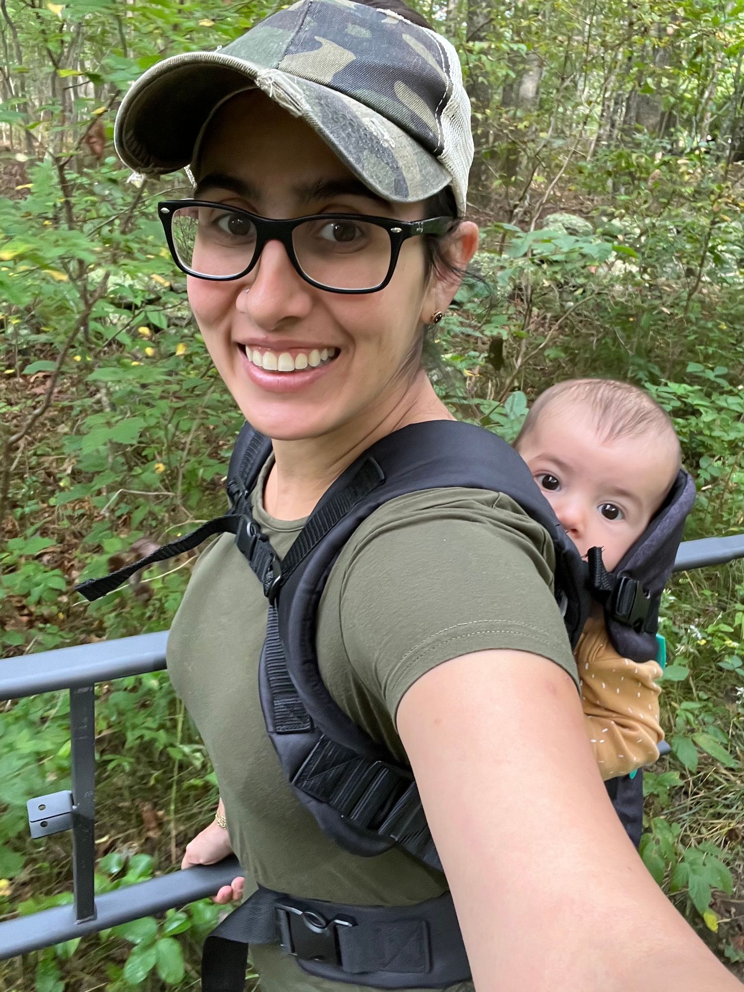 A hiker wearing glasses and a baseball cap carries a baby in a backpack carrier on a woodland trail.
