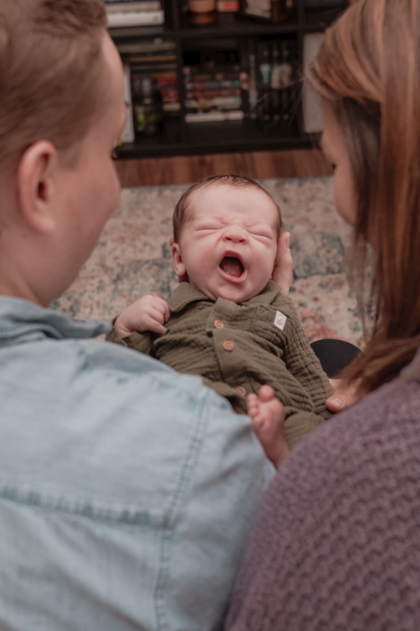 A newborn baby yawns while being held by adults in denim and purple sweaters.
