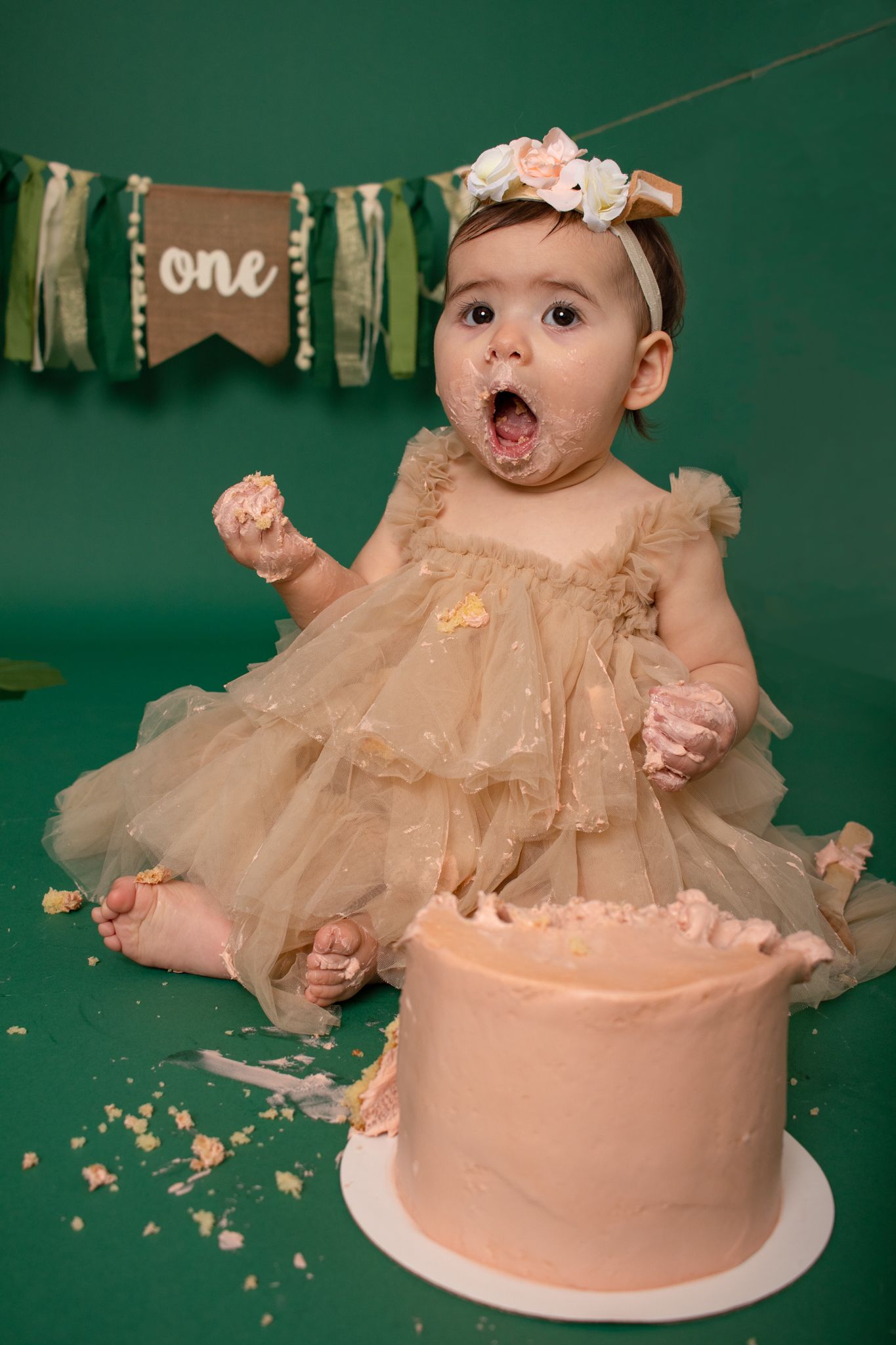 Cake smash session with beige dress against green backdrop with 'one' banner.