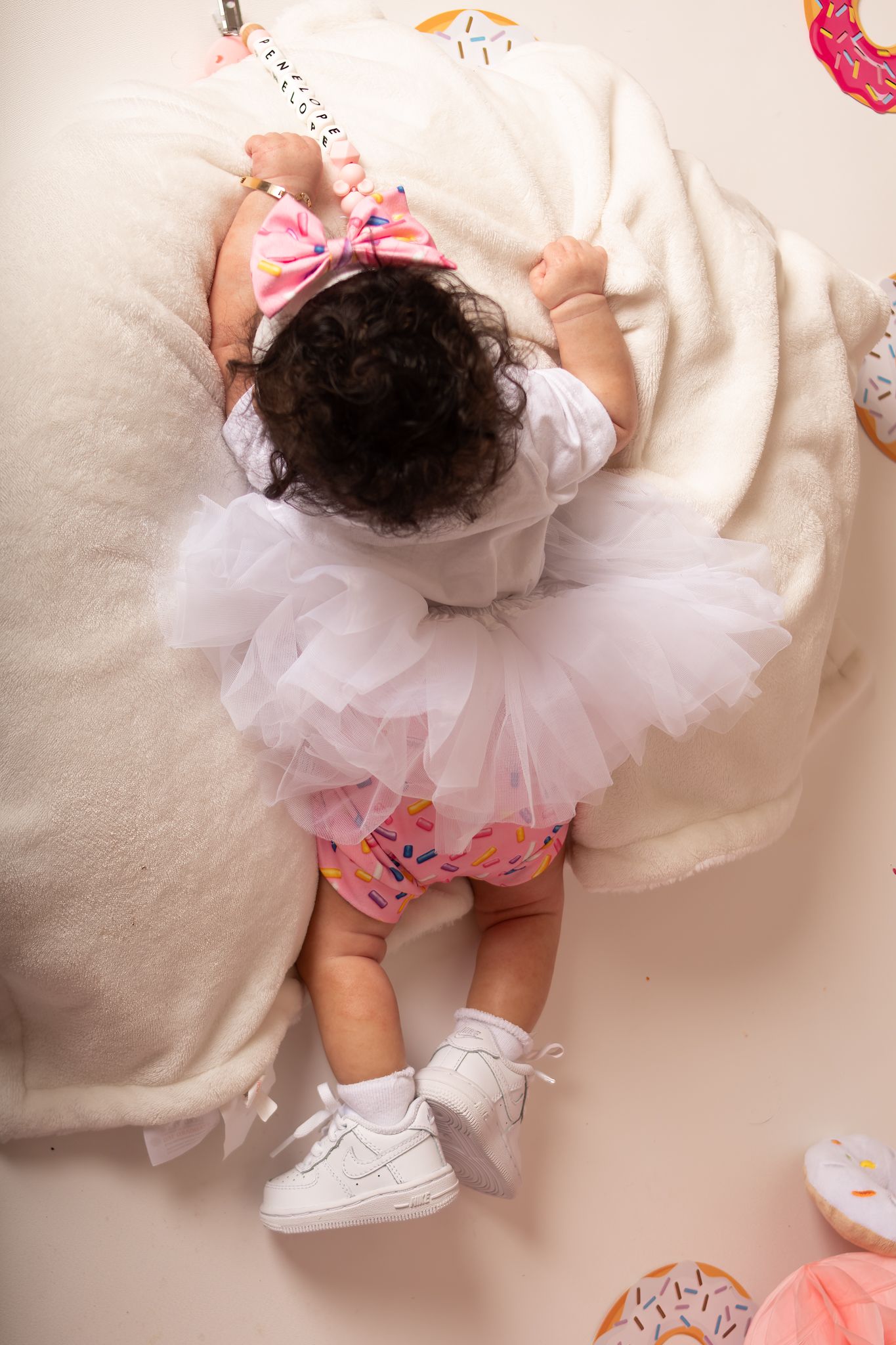 Overhead view of baby wearing white tutu and shoes lying on soft pillows with pink accessories.