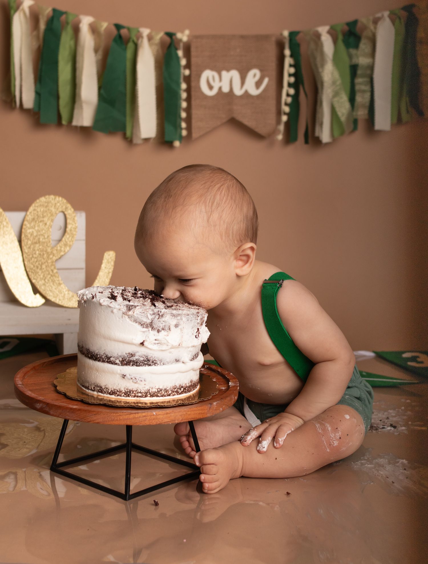 Baby in green suspenders leaning over a white cake at first birthday photoshoot.