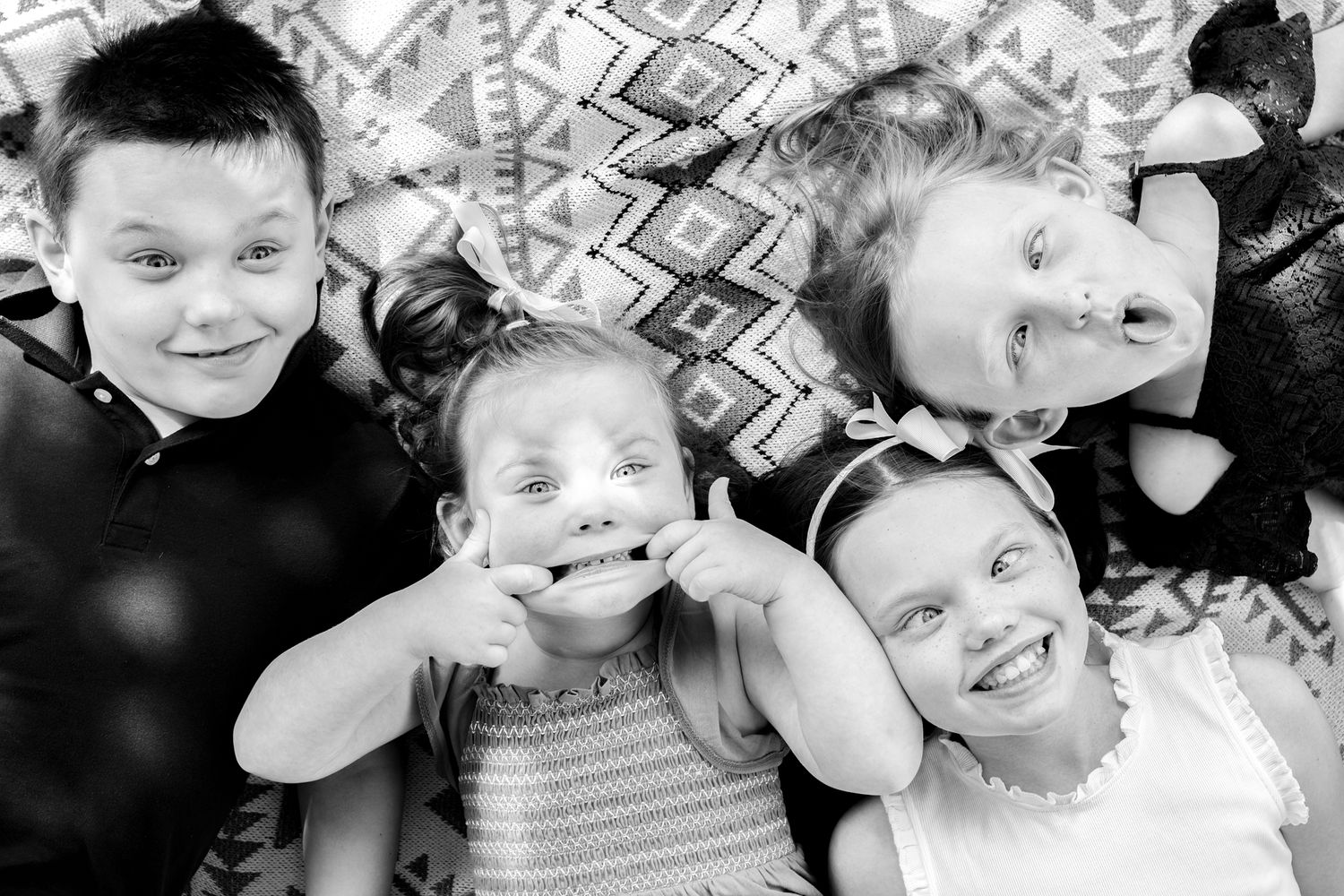 Black and white overhead shot of children making silly faces while lying on a patterned blanket.