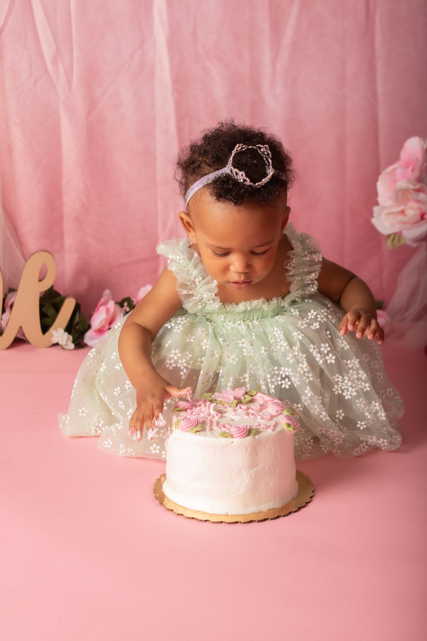 Baby in lace dress celebrates first birthday with pink cake against pink backdrop.