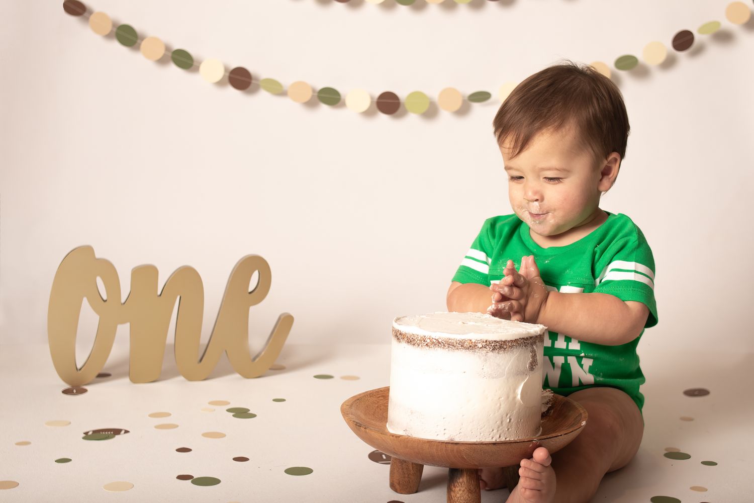 One year old cake smash photo with white cake and gold 'one' text decoration against neutral background.