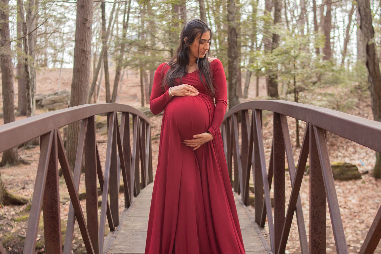 Expectant mother in flowing red dress poses on wooden bridge in Smithfield RI forest.