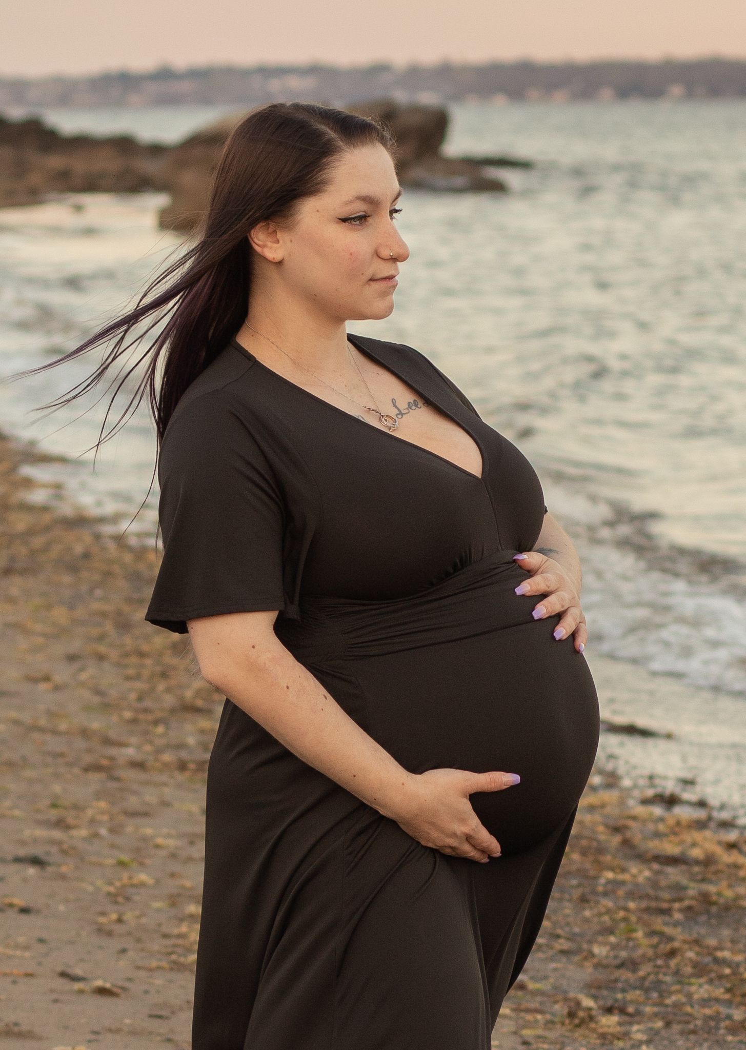 A maternity portrait captures an expectant mother in a black dress against a coastal backdrop.