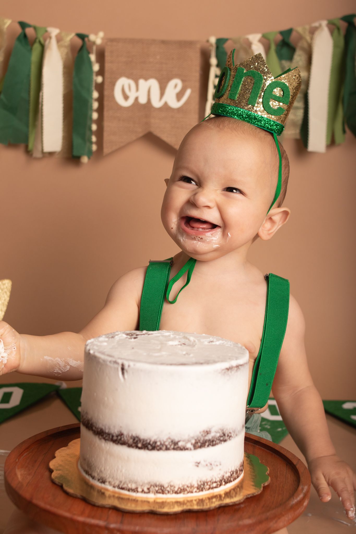 First birthday celebration with cake, green decorations and smiling baby wearing crown.