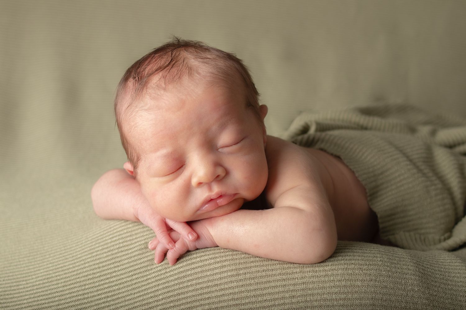 Newborn baby sleeping soundly on soft sage green blanket in natural light.