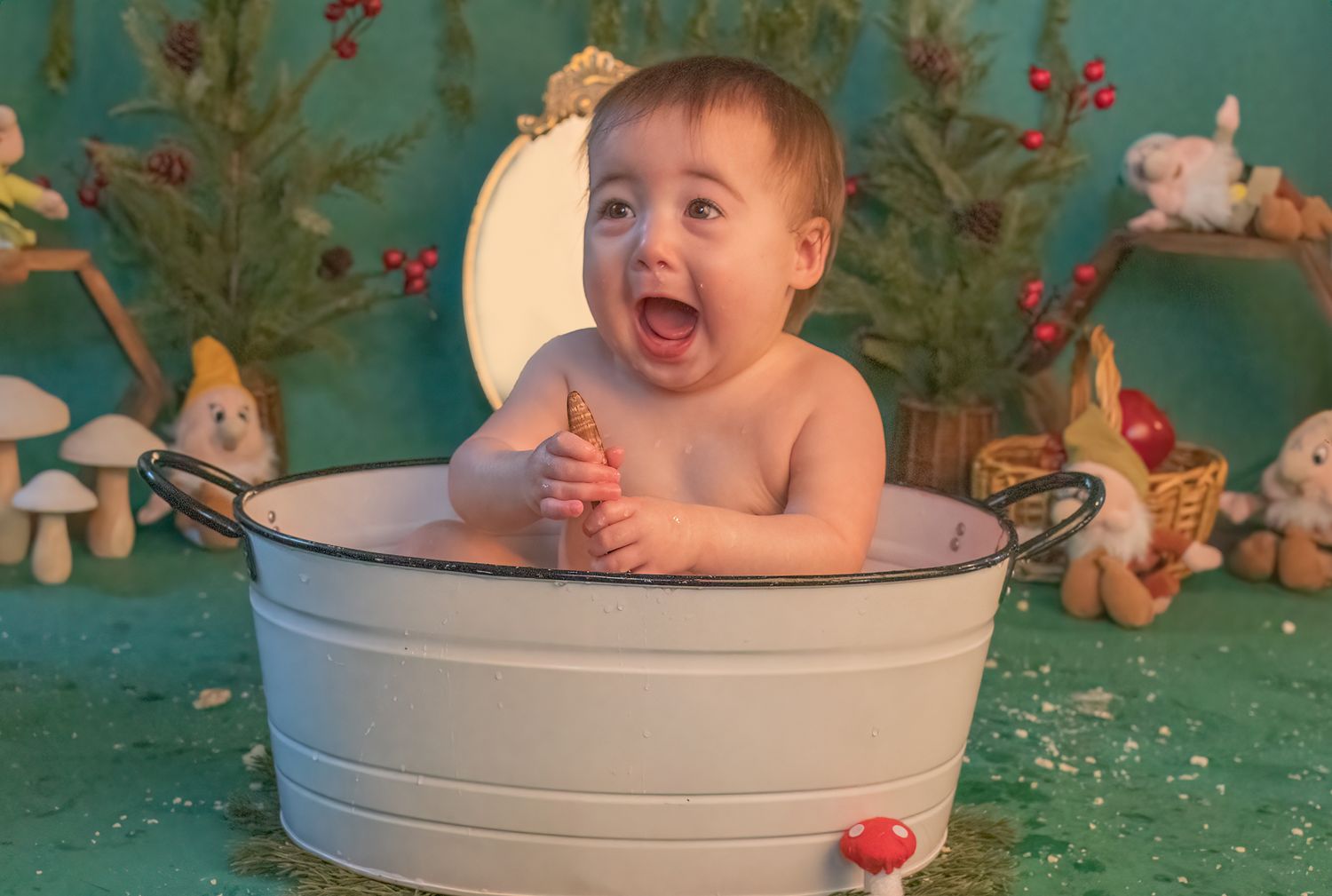 Happy baby bathing in white metal tub with holiday decorations in background.
