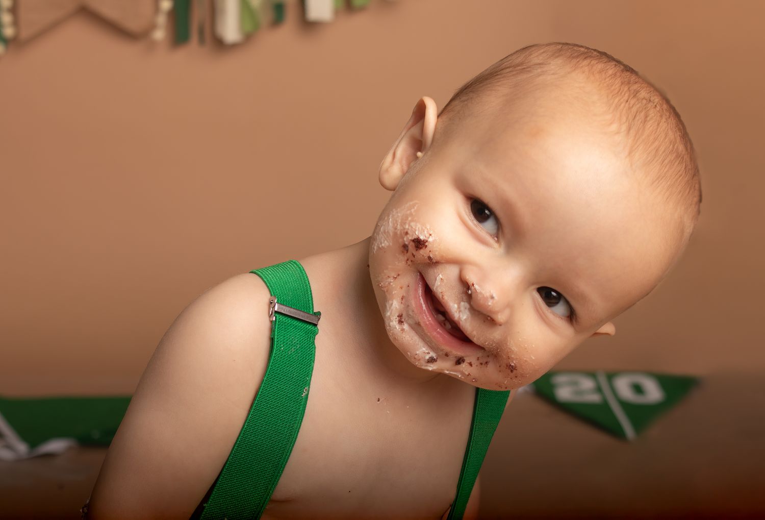 Close up of smiling baby with cake frosting on face wearing green suspenders.