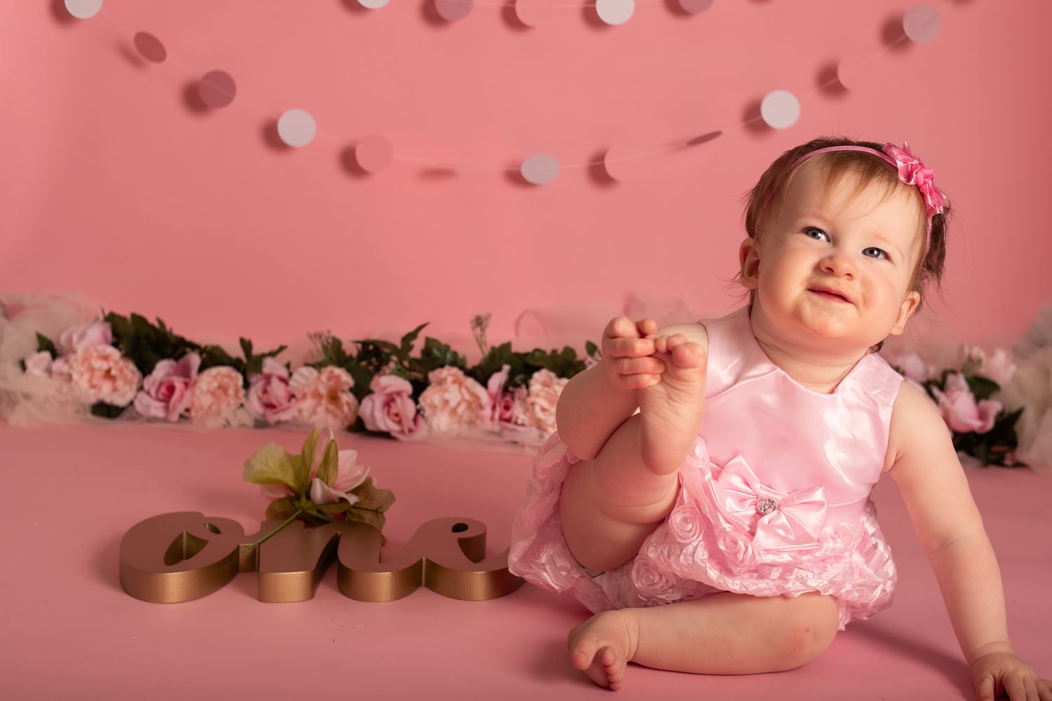 Baby in pink dress sitting against floral backdrop with string lights.