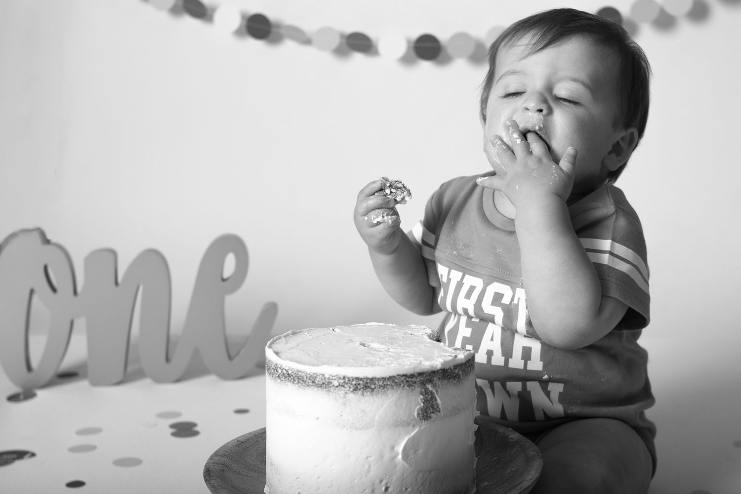 Black and white photo of a first birthday celebration with cake and decorative 'one' sign.