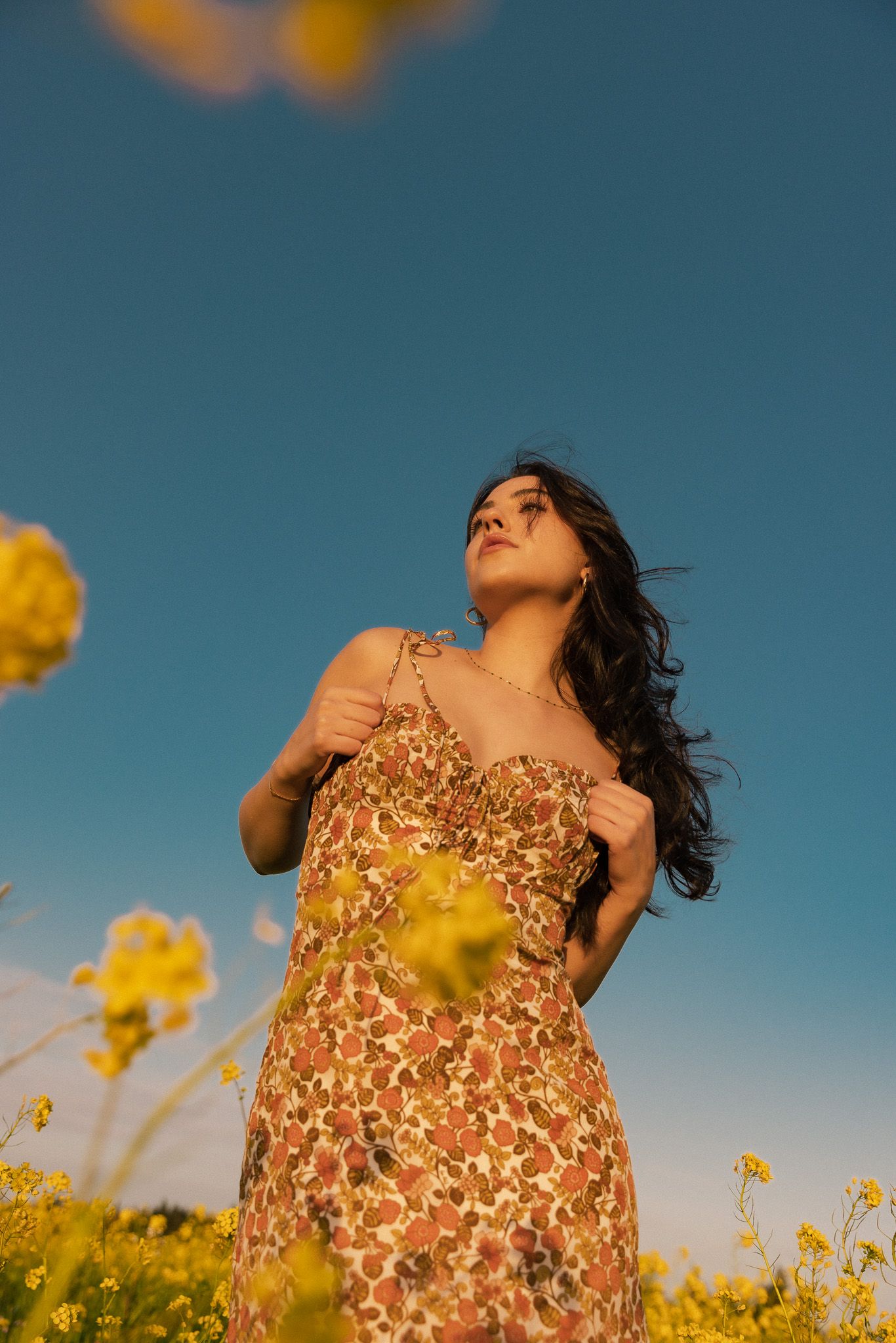 Someone in a floral dress poses among yellow wildflowers against a vibrant blue sky during golden hour.