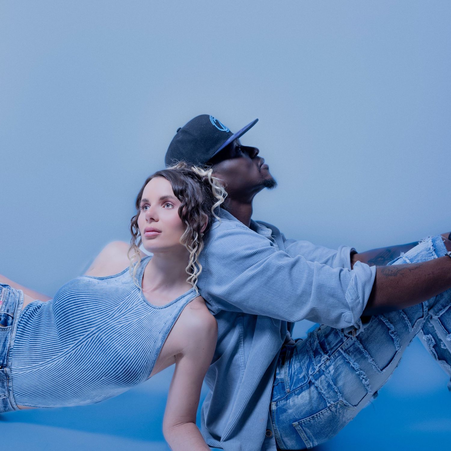 Two models pose back-to-back against a blue background wearing denim and casual clothing during a fashion shoot.