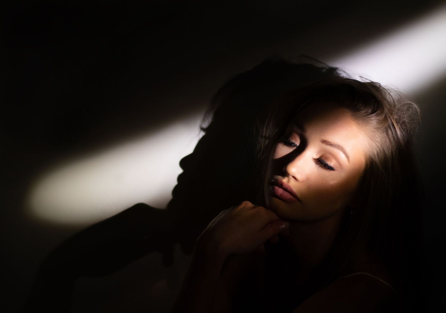 Dramatic portrait in shadow and light with bold diagonal beam illuminating soft features against dark background.