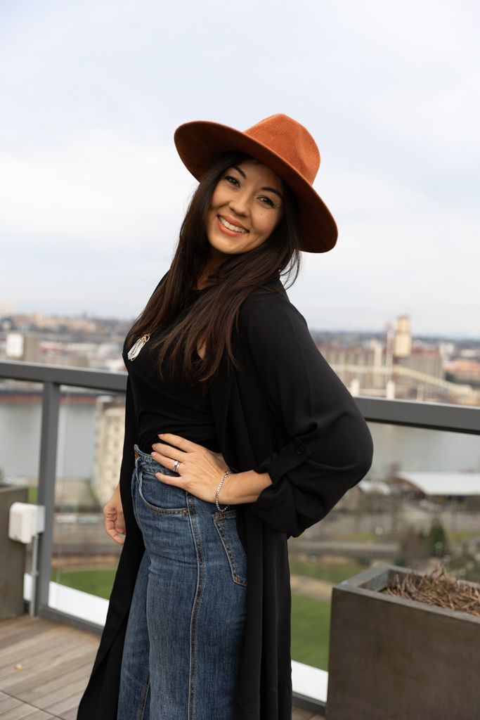 Stylish woman in brown hat, black cardigan and jeans poses outdoors on rooftop terrace with cityscape in background.