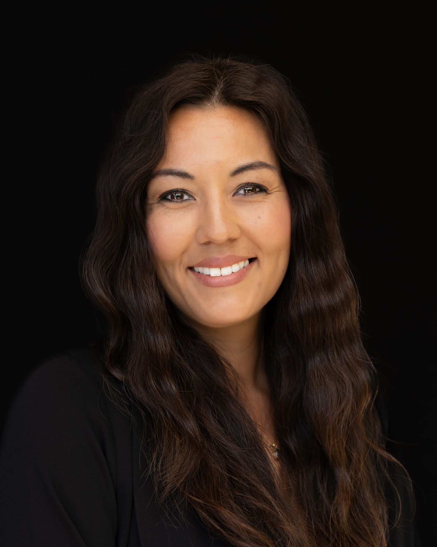 Professional headshot portrait of someone with long dark wavy hair and a warm smile wearing black against dark background.