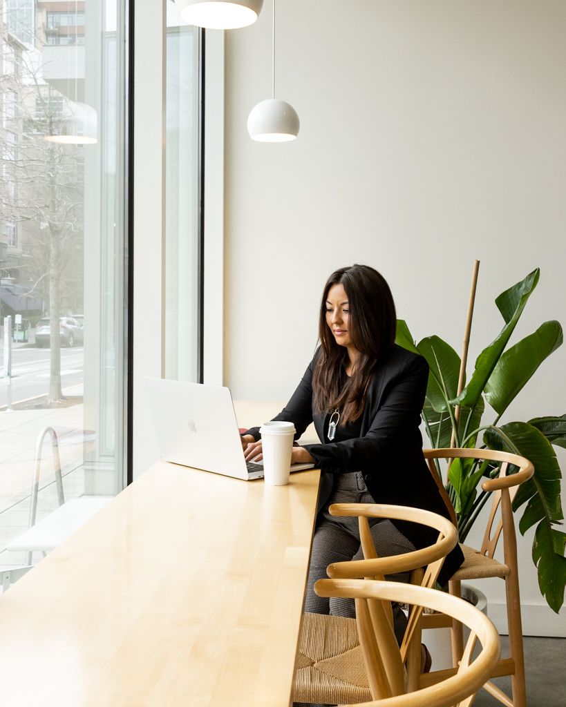 Professional working on laptop at wooden desk by window with natural light and houseplant in modern minimalist office space.