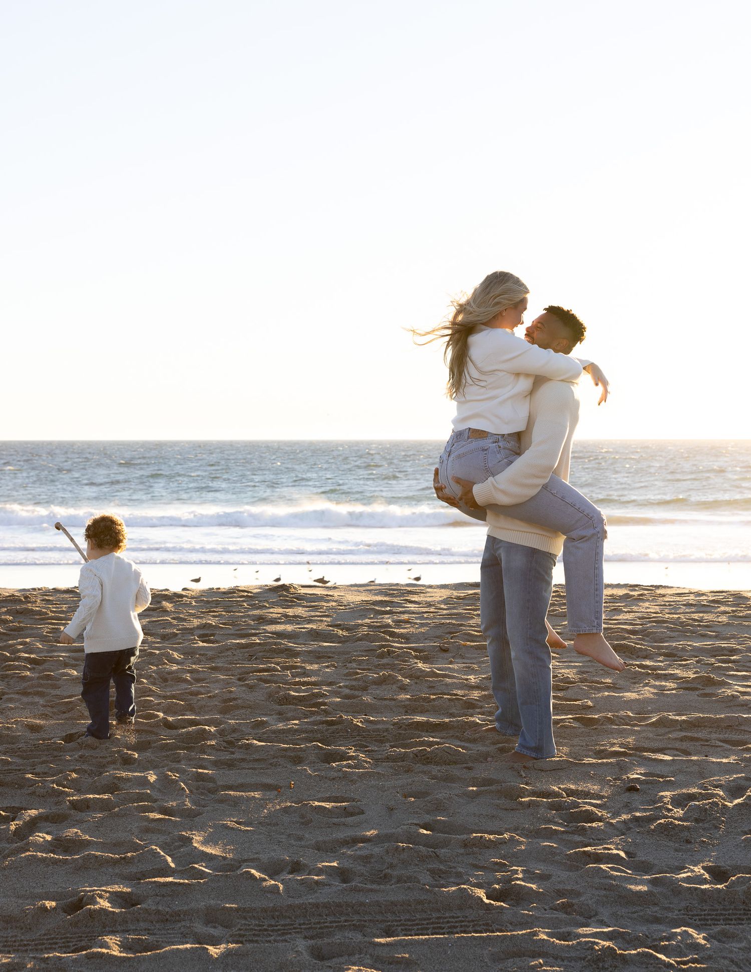 Embracing couple in casual attire and small child standing on sandy beach during sunset with ocean in background.