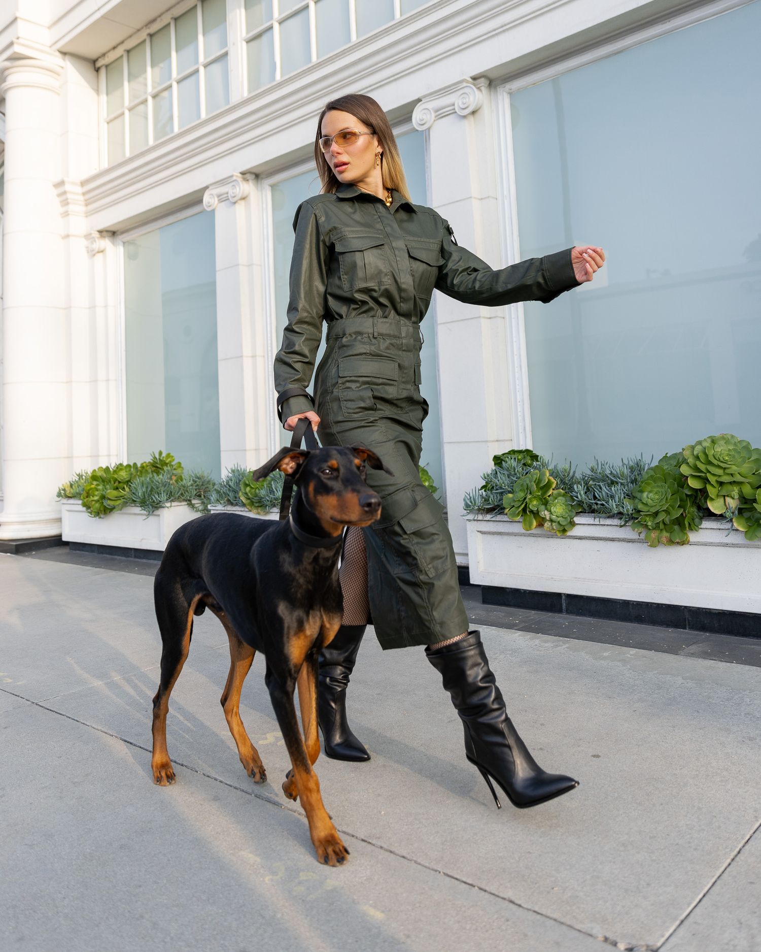 Stylish military-inspired olive jumpsuit paired with tall black boots and a well-groomed Doberman dog standing on a sidewalk.