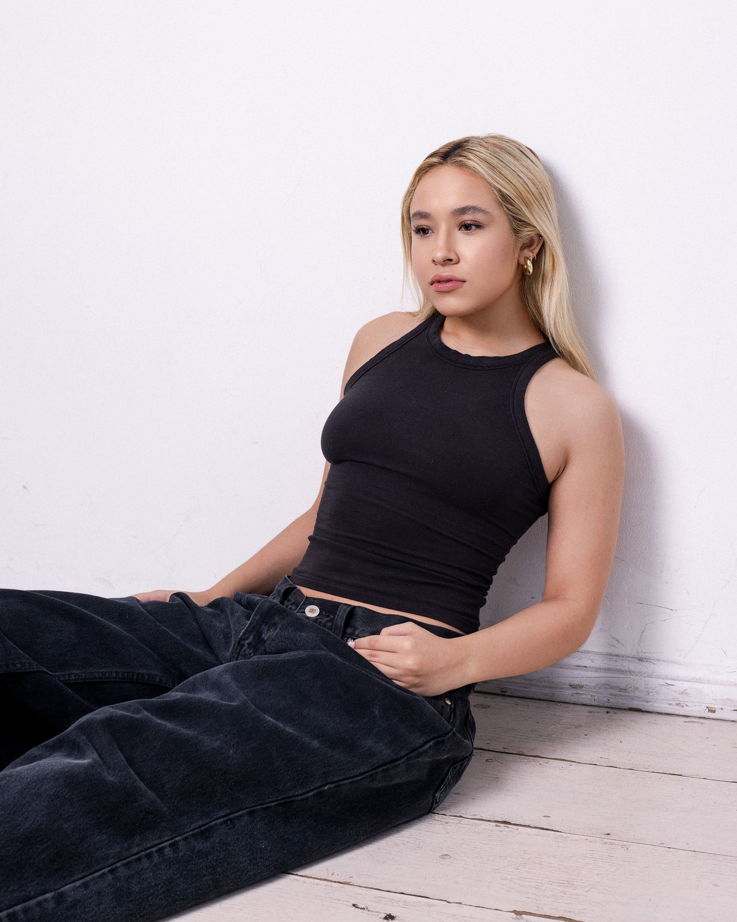 A person in a black crop top and dark jeans sits on a white wooden floor against a plain wall.