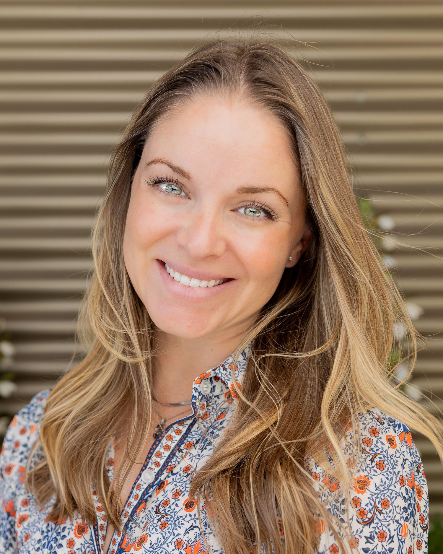 Professional headshot of someone with long blonde hair and bright blue eyes wearing a floral print blouse against a striped background.
